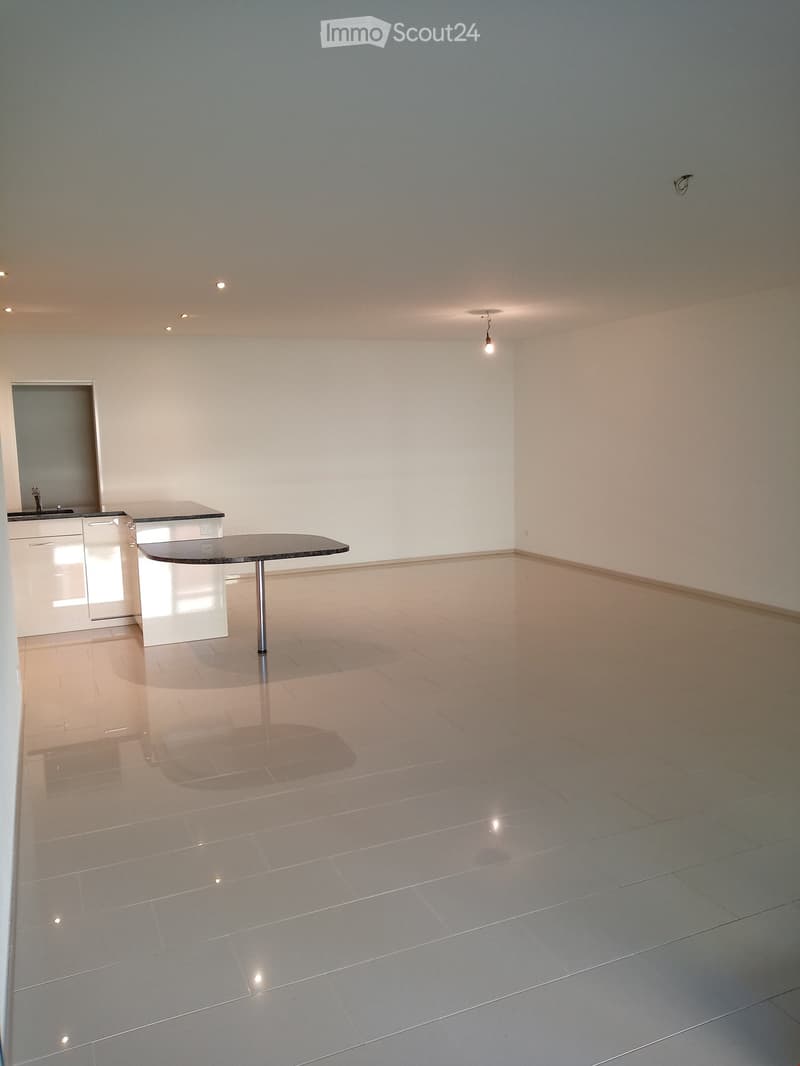 Empty room with white tiles, a round table, a kitchen area with a sink, and a ceiling with lights.