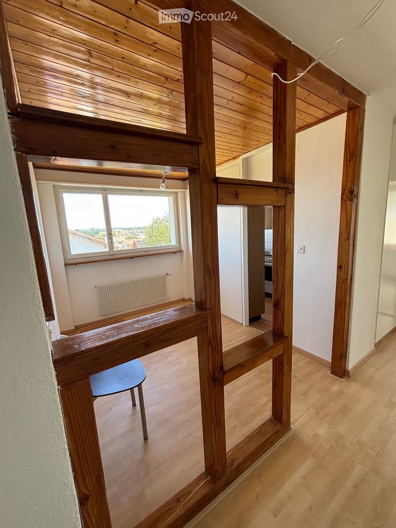 Wooden staircase with a blue chair on the landing, leading to an upper level. The hallway has light wooden flooring, white walls, and a window with a view of the outside.
