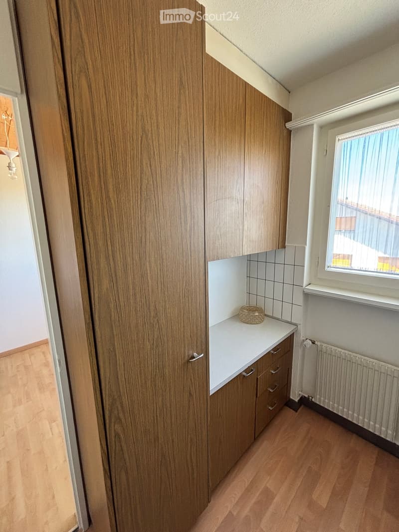 A small, furnished room with wooden cabinets, a countertop, a drawer, a radiator, and a wicker basket on the counter. The room has wooden flooring and a window with blinds.
