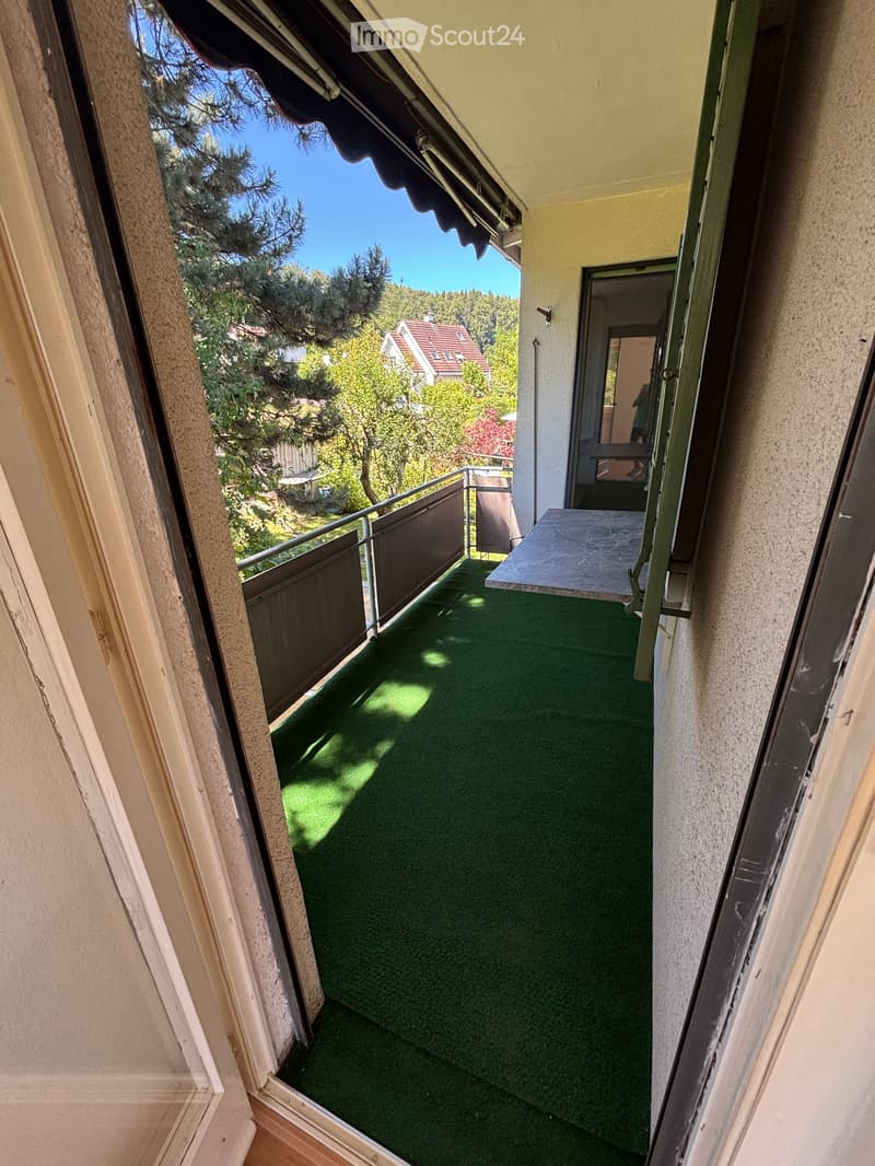 Balcony with green artificial turf, metal railing, and a small table, overlooking a tree-lined area with houses in the background.