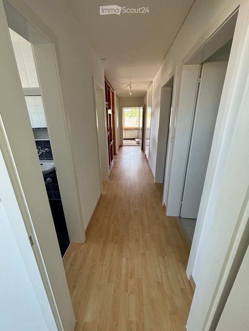 Long, narrow hallway with light wooden flooring and white walls, featuring several doors and a window at the end.