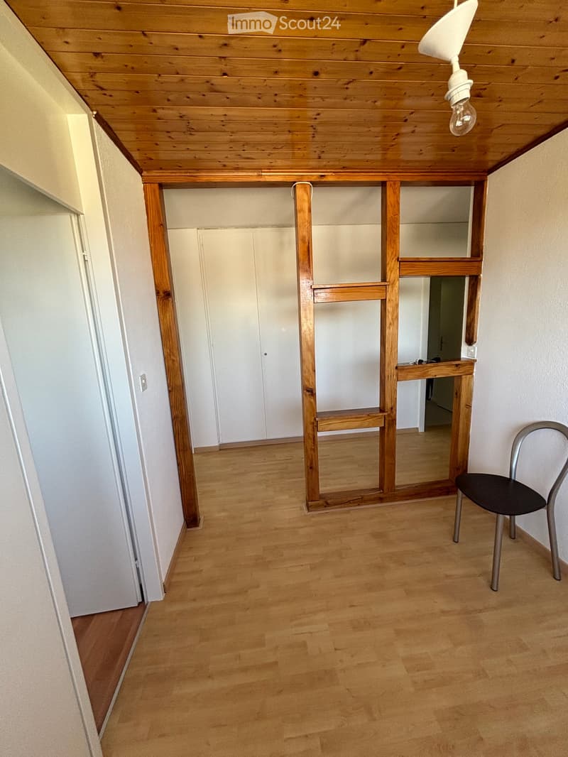 A small, unfurnished room with wooden flooring, white walls, and a single chair near the corner. The room features a wooden shelf structure and a closed white cabinet.