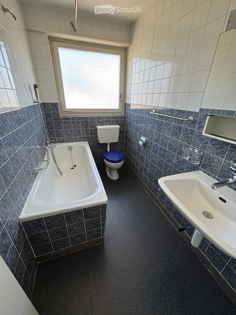 Bathroom with a bathtub, toilet, sink, and mirror. The walls are tiled in grey, and the floor is dark grey.
