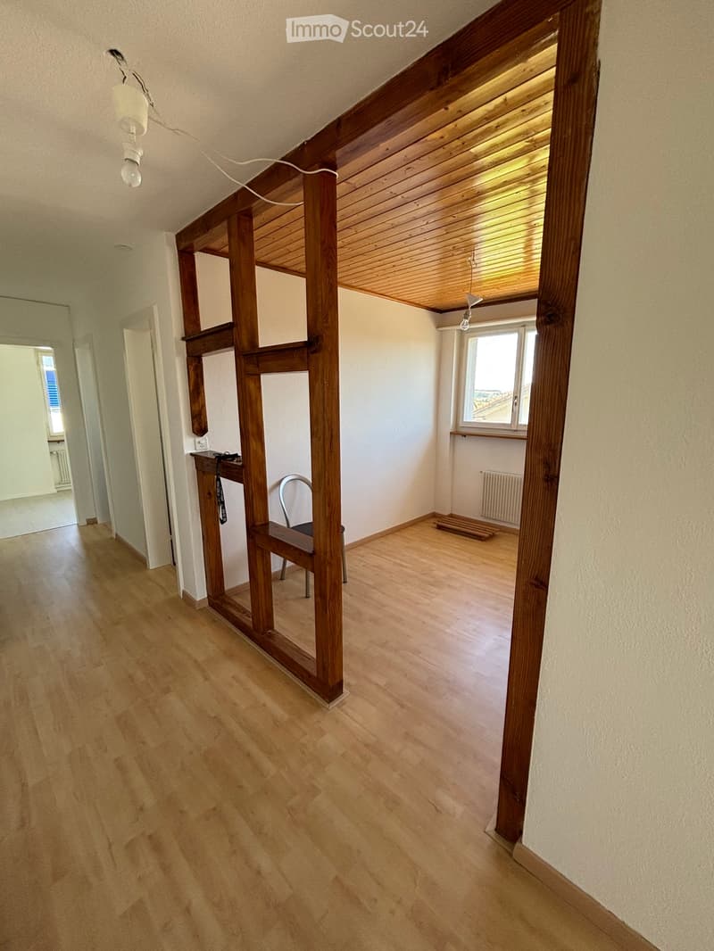 Empty room with wooden flooring, white walls, and a wooden staircase leading to an upper level. There is a small table with a chair, a window with blinds, and a radiator visible.