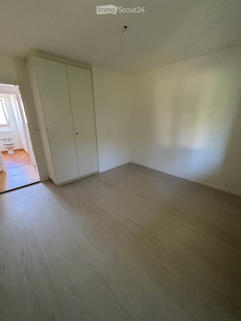 Empty room with light-colored wooden flooring and white walls. A built-in closet is visible on one wall, and an open doorway leads to another room with a toilet.