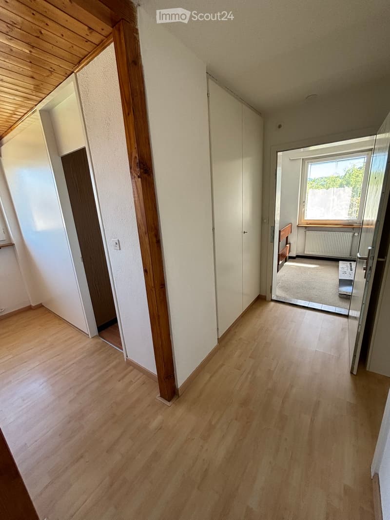 Empty hallway with wooden flooring, white walls, and a large window at the end, leading to another room.