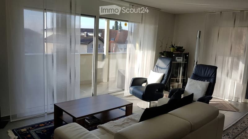 Modern living room with large windows, a sofa, coffee table, two chairs, and a shelf with decorative items.