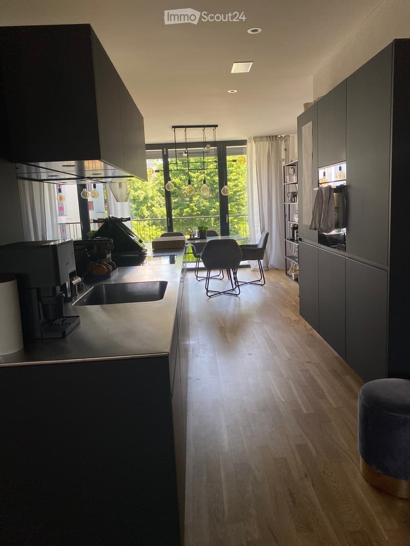 Modern kitchen with dark cabinets, light wooden flooring, and a dining area with chairs and a table. Features pendant lights, a coffee machine, and a sink.