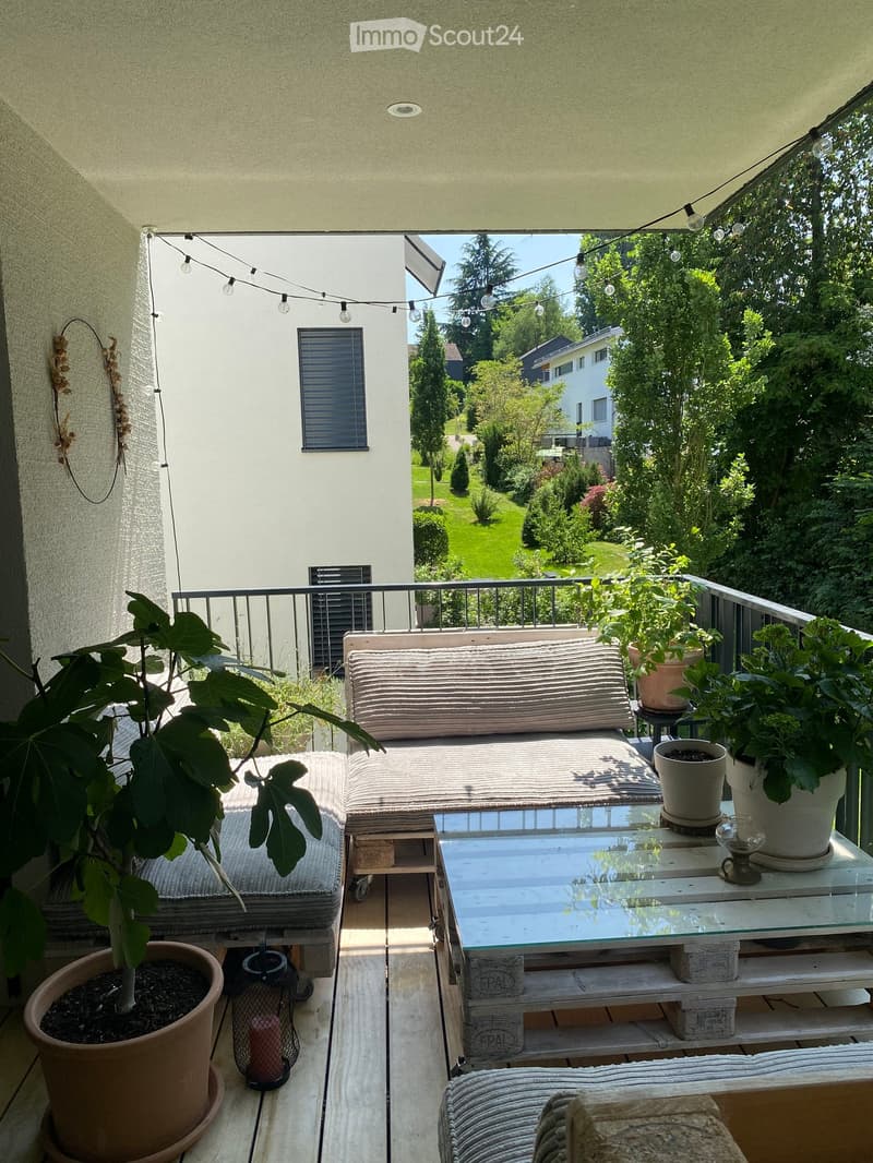 Balcony with a sofa, coffee table, and several potted plants, surrounded by greenery and a view of a garden.