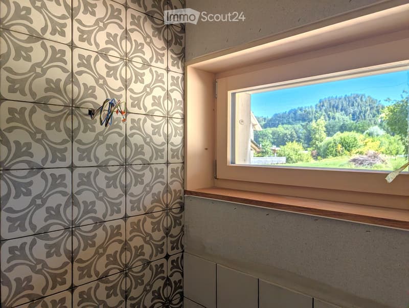 Empty room with patterned tiles on one wall, a window with a scenic mountain view, and a wooden shelf below the window.