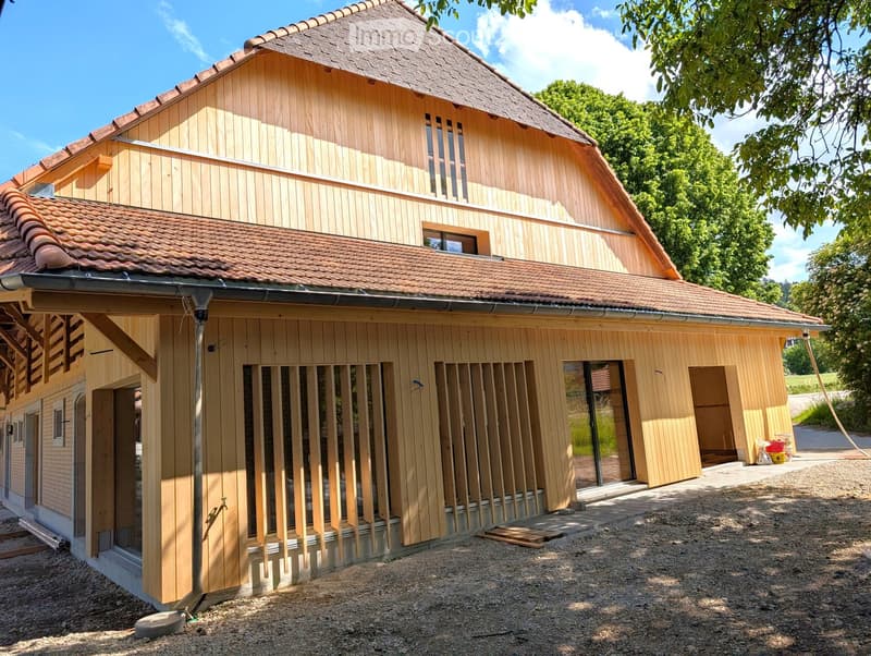 Two-story wooden house with a tiled roof, large windows, and a partially constructed exterior. The house features a mix of wooden and brick walls, with visible construction materials and tools near the entrance.