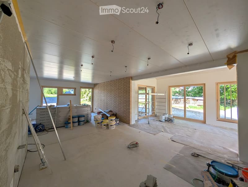 Empty room with exposed wooden beams, white walls, and large windows. Construction materials and tools are scattered around, indicating ongoing renovation.