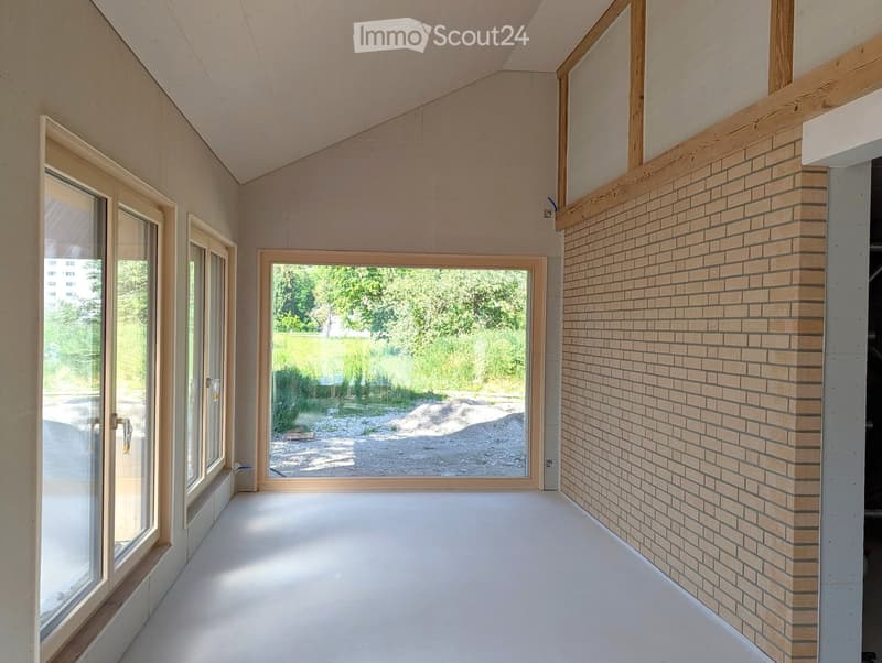 Empty room with white walls, wooden floor, large windows, and a brick accent wall.