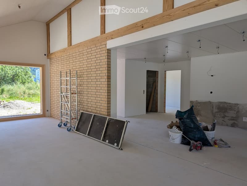 Empty room with exposed wooden beams, brick wall, large window, and construction materials including a ladder, buckets, and tools.