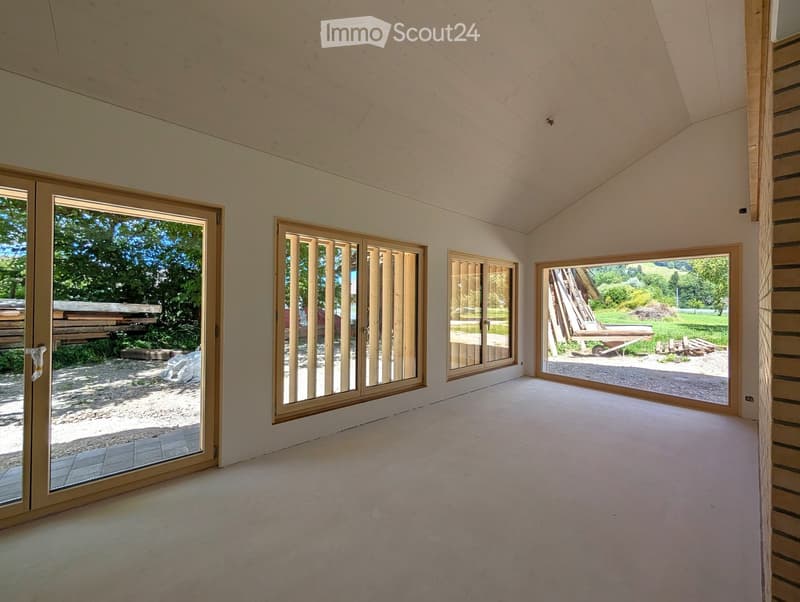 Empty room with large windows and wooden beams, showing natural light and an outdoor view.
