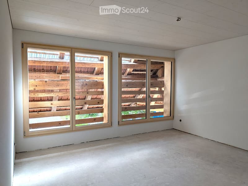 Empty room with concrete floor, white walls, and large windows with wooden frames showing a wooden deck outside.