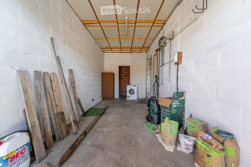 Empty room with white brick walls, exposed wooden beams on the ceiling, and various construction materials and tools scattered around, including a washing machine, wooden planks, and bags of cement.