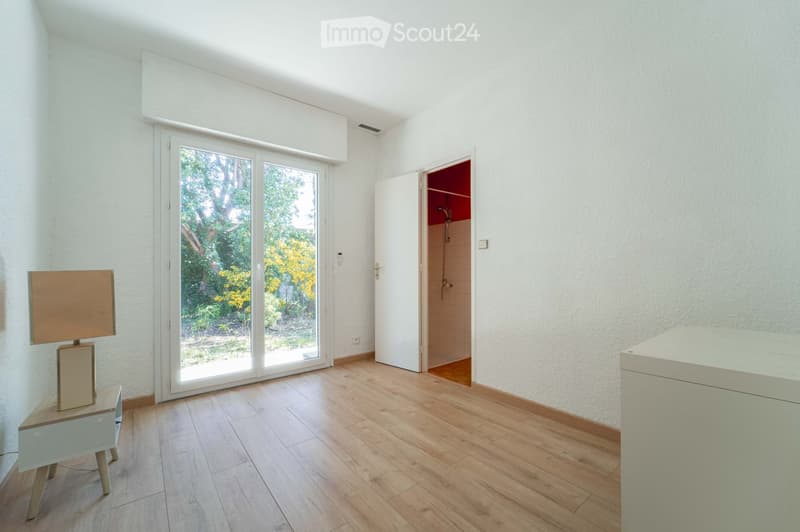 Bright, minimalist room with wooden flooring, large sliding glass doors leading to a garden, a small table with a lamp, and an open door to a bathroom.