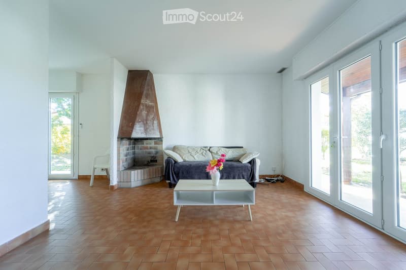 Bright living room with wooden flooring, white walls, a fireplace, a sofa, a coffee table, and a chair near the window.