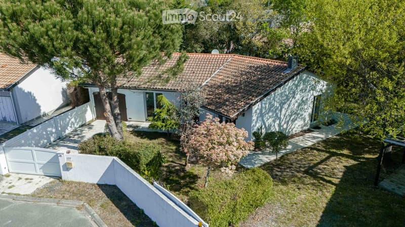 Aerial view of a two-story house with a tiled roof, surrounded by trees and a well-maintained garden. The house has a white exterior with multiple windows and a small porch area.