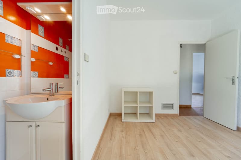 Modern bathroom with white walls, wooden flooring, double sink, and built-in cabinetry. Features a bright orange accent wall with decorative tiles and a large mirror above the sink.
