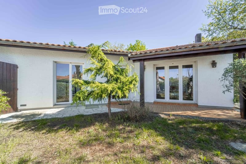 Single-story house with white walls, tiled roof, large glass doors, and a small garden area with a tree and pathway.