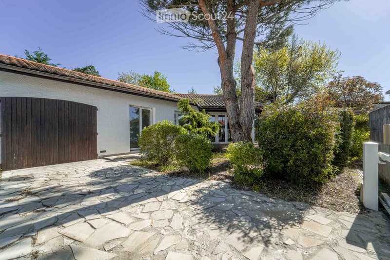 A modern house with a tiled roof, white exterior, and large wooden garage door. The entrance features a stone pathway leading to the house, surrounded by greenery and trees.