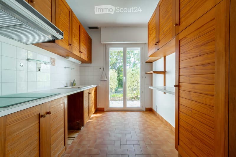 Bright kitchen with wooden cabinets, tiled walls, and a central island. Features a sink, faucet, and range hood, with a sliding glass door leading to a garden.
