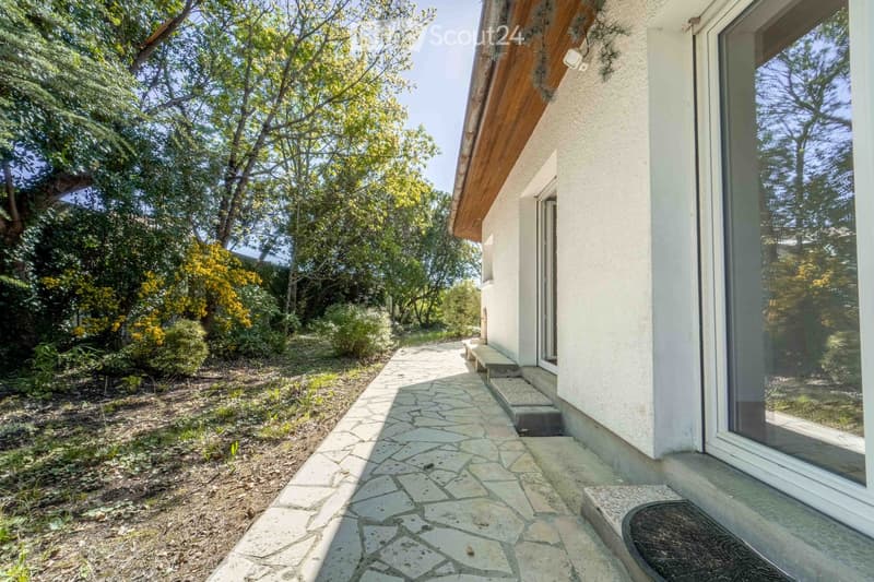 A pathway leads to a house with a modern design, featuring a raised ground floor and a wooden roofline. The surrounding area is lush with trees and greenery.
