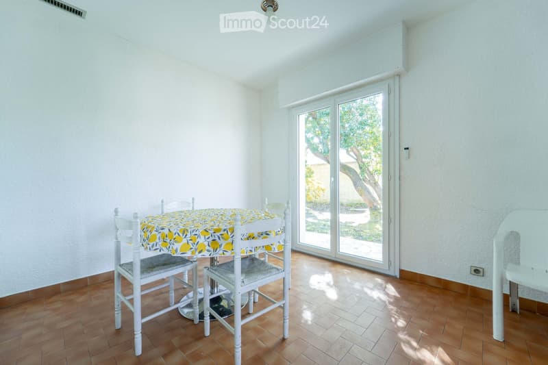 Bright dining room with wooden flooring, white walls, and a large sliding glass door leading to a garden. A round table with a lemon-patterned tablecloth and four matching chairs are present.