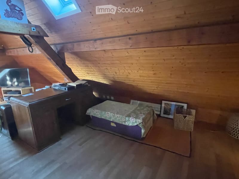 A cozy attic room with wooden walls and a slanted ceiling, featuring a refrigerator, a small table with a patterned cloth, a chair, a TV screen, and a cabinet.