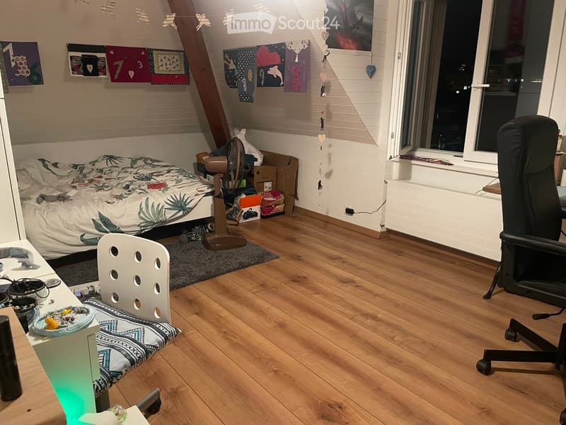 Cozy bedroom with wooden flooring, a bed with a colorful blanket, a chair, a fan, a small table with items, and a window with a view of the night sky.