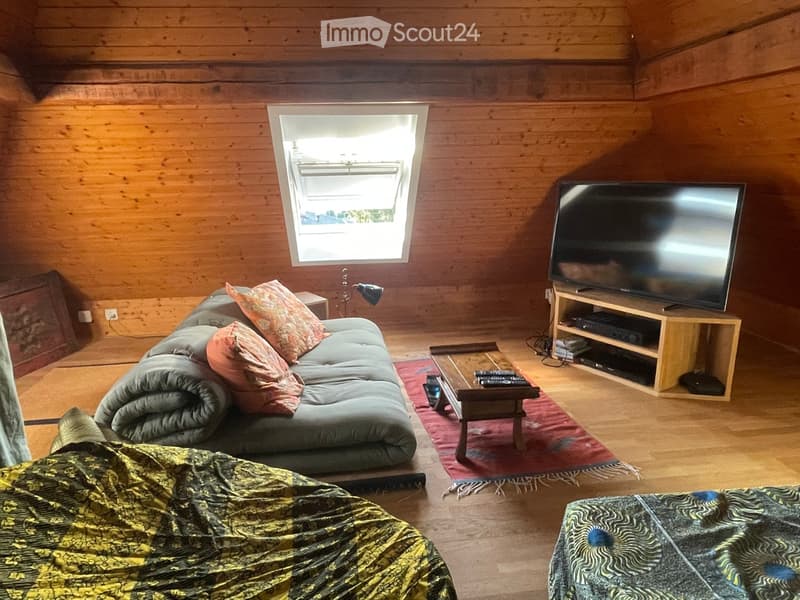 A cozy living room with wooden walls and floors, featuring a grey sofa with colorful pillows, a coffee table with remote controls, a television on a wooden stand, and a small side table with a lamp.
