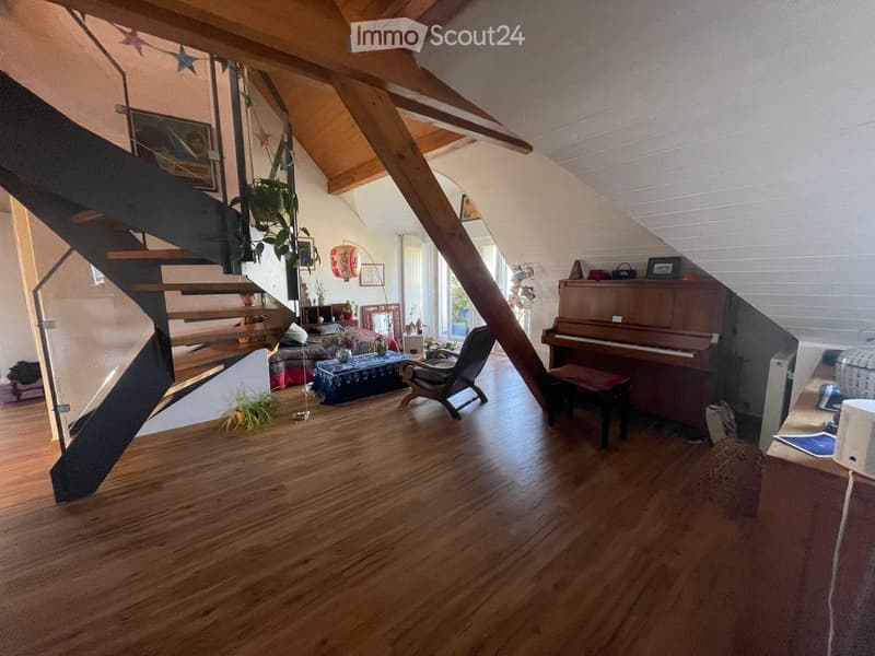 A spacious, well-lit living area with wooden flooring, a grand piano, a staircase leading to an upper level, and several seating options including a chair and small tables.