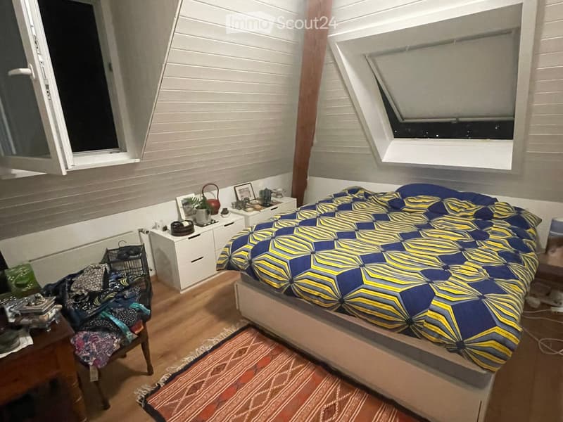 Cozy bedroom with a bed covered in a blue and yellow patterned duvet, a nightstand with a plant and decorative items, a chair with a colorful cushion, and a small table with various items.