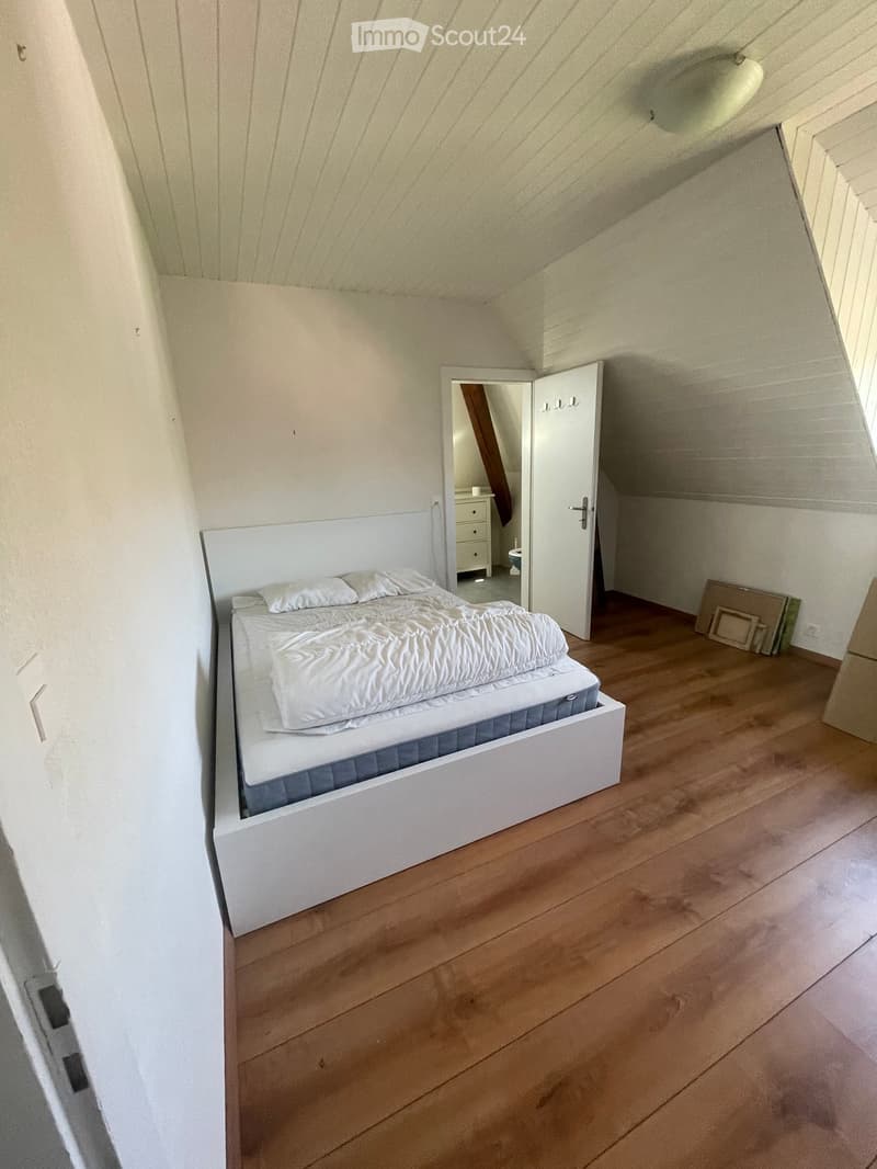 Bright attic bedroom with wooden flooring, white walls, and a large bed with white bedding and pillows. A nightstand and a small stool are visible in the adjacent room.