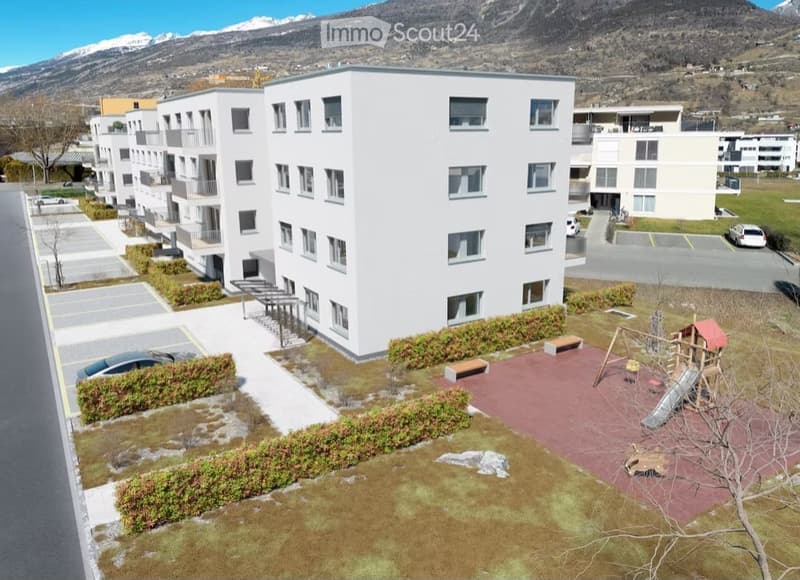 Modern multi-story residential building with white facade and multiple windows, surrounded by a landscaped area with playground equipment, benches, and small trees.