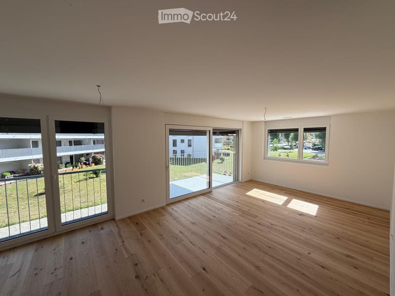 Empty room with wooden flooring, large windows, and sliding glass doors leading to a balcony area.