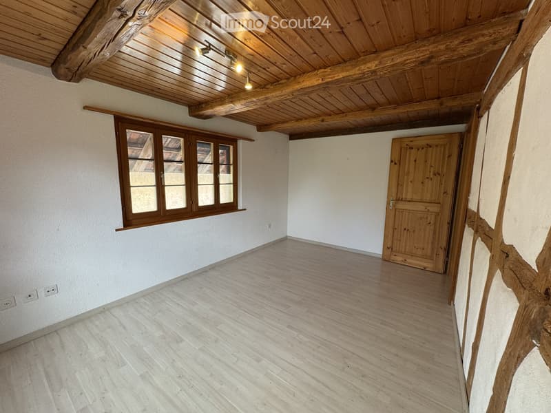 Empty room with wooden floor, white walls, wooden ceiling beams, and a large window with wooden frame. No furniture or equipment.