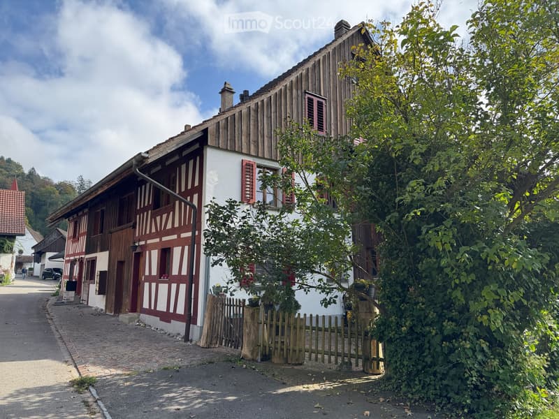 Traditional half-timbered house with white walls, red shutters, and wooden beams, surrounded by greenery and a wooden fence.