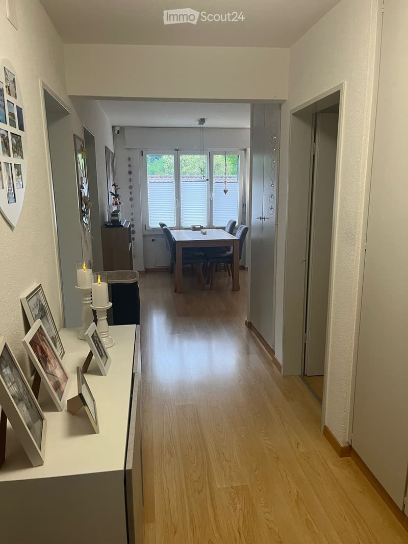 Spacious hallway with light wooden flooring, white walls, and framed pictures on a console table. The hallway leads to a dining area with a table and chairs, large windows, and a view of greenery outside.