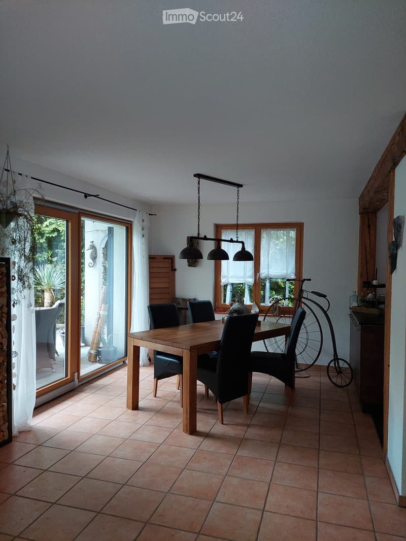 Dining room with a wooden table, six chairs, and a hanging lamp. Large windows and sliding glass doors lead to an outdoor area.