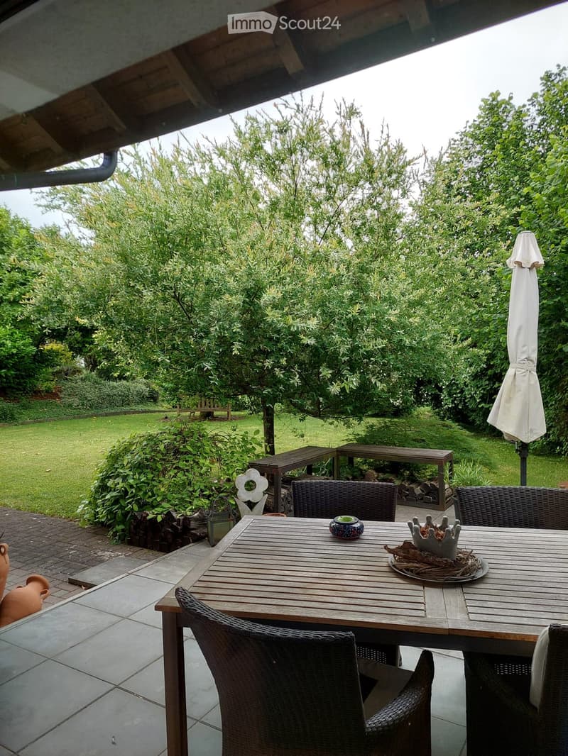 Outdoor patio with wooden table, wicker chairs, umbrella, and decorative items, surrounded by greenery and a lawn.