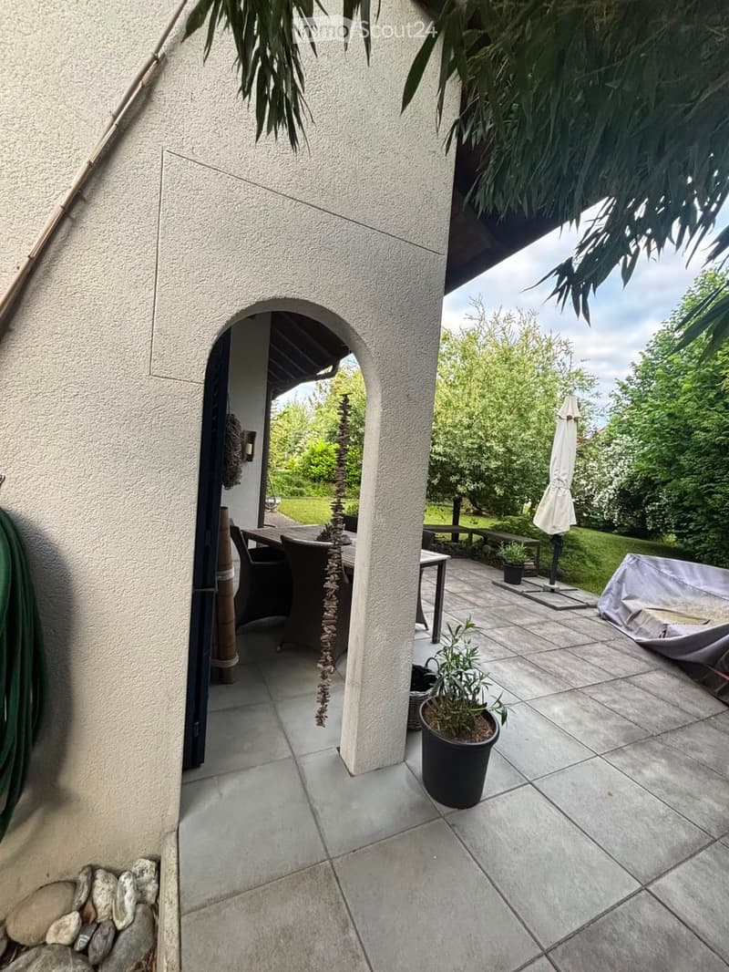 Outdoor patio area with tiled flooring, a table and chairs, an umbrella, a bench, and potted plants.