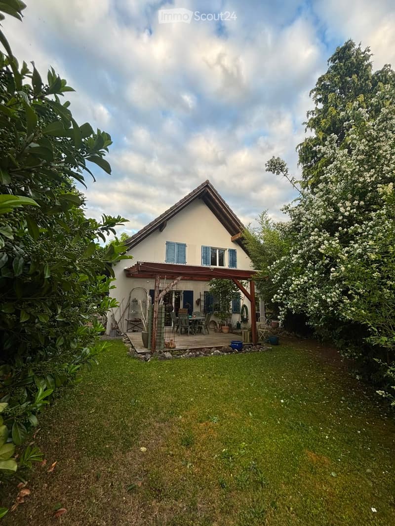 A two-story house with a pitched roof, light-colored exterior, and blue shutters. The house features a covered patio area with outdoor furniture and is surrounded by a well-maintained garden with various plants and trees.
