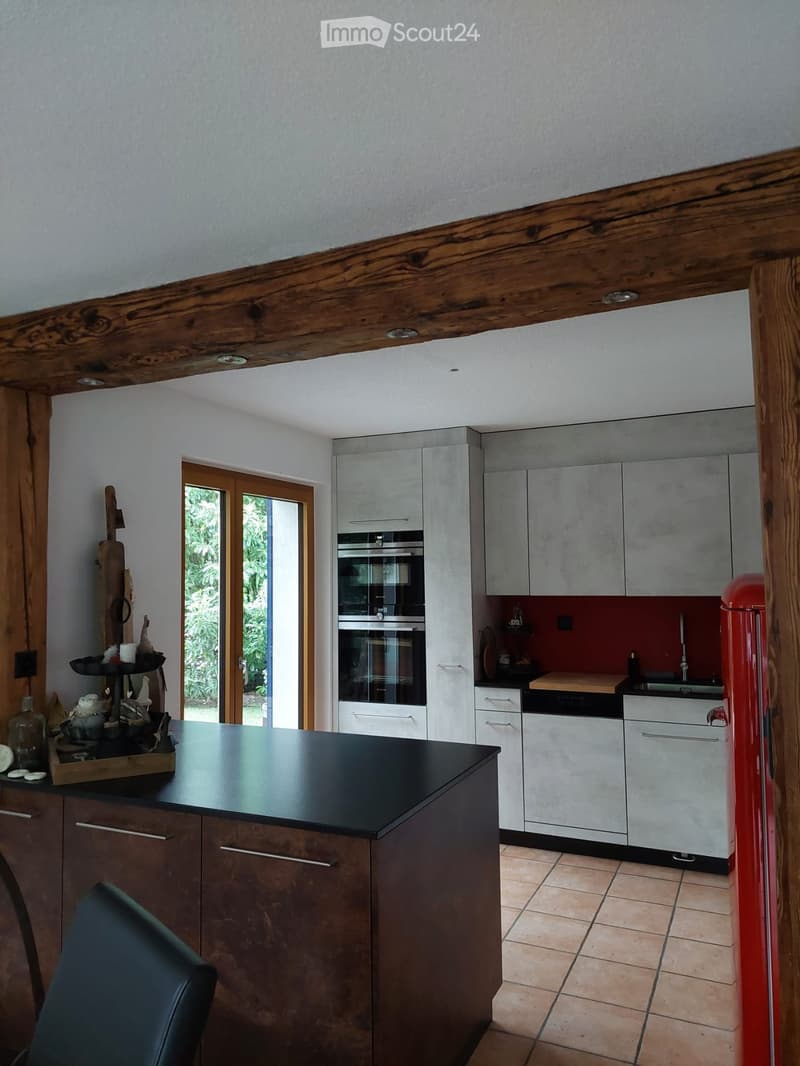 Modern kitchen with grey cabinets, stainless steel oven, red refrigerator, and a black countertop. Wooden beams on the ceiling and a tiled floor.
