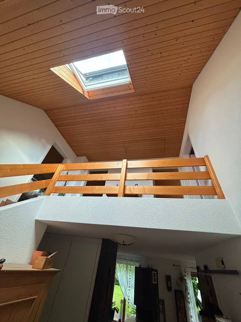 A wooden balcony with a railing and a skylight above, overlooking a lower level with a wooden floor and white walls.