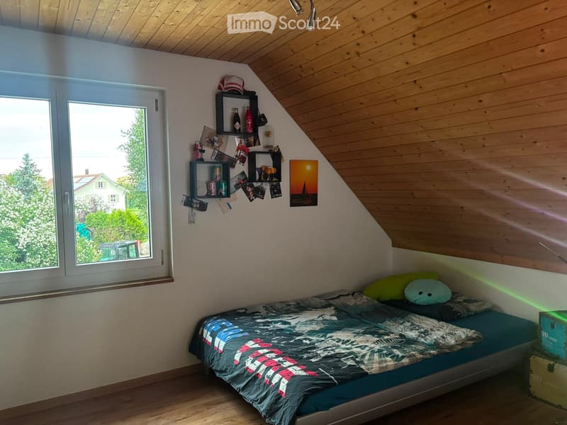 Cozy attic bedroom with wooden slanted ceilings, large window offering a view of greenery, bed with colorful bedding, shelves with various items, and a picture frame on the wall.