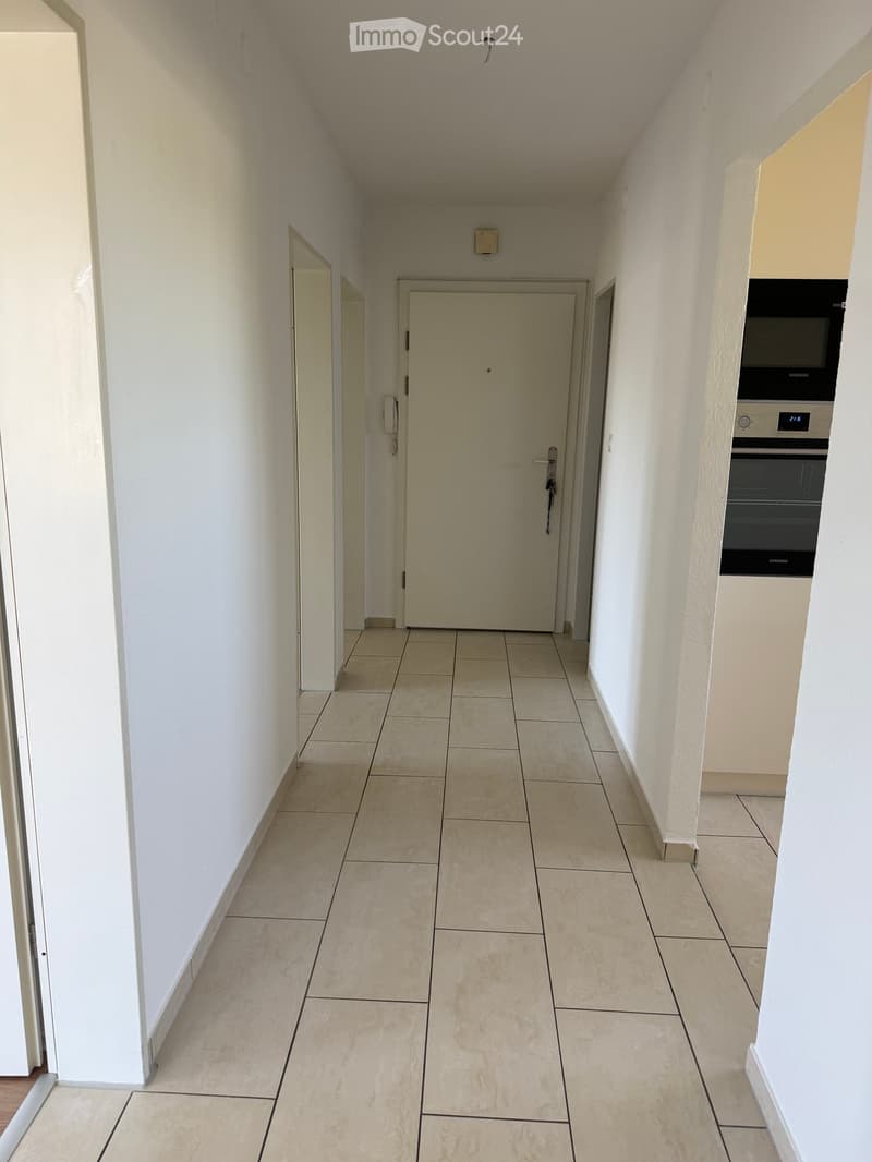 Empty hallway with tiled floor, white walls, and doors leading to other rooms. A small section of a kitchen with a built-in oven is visible on the right side.
