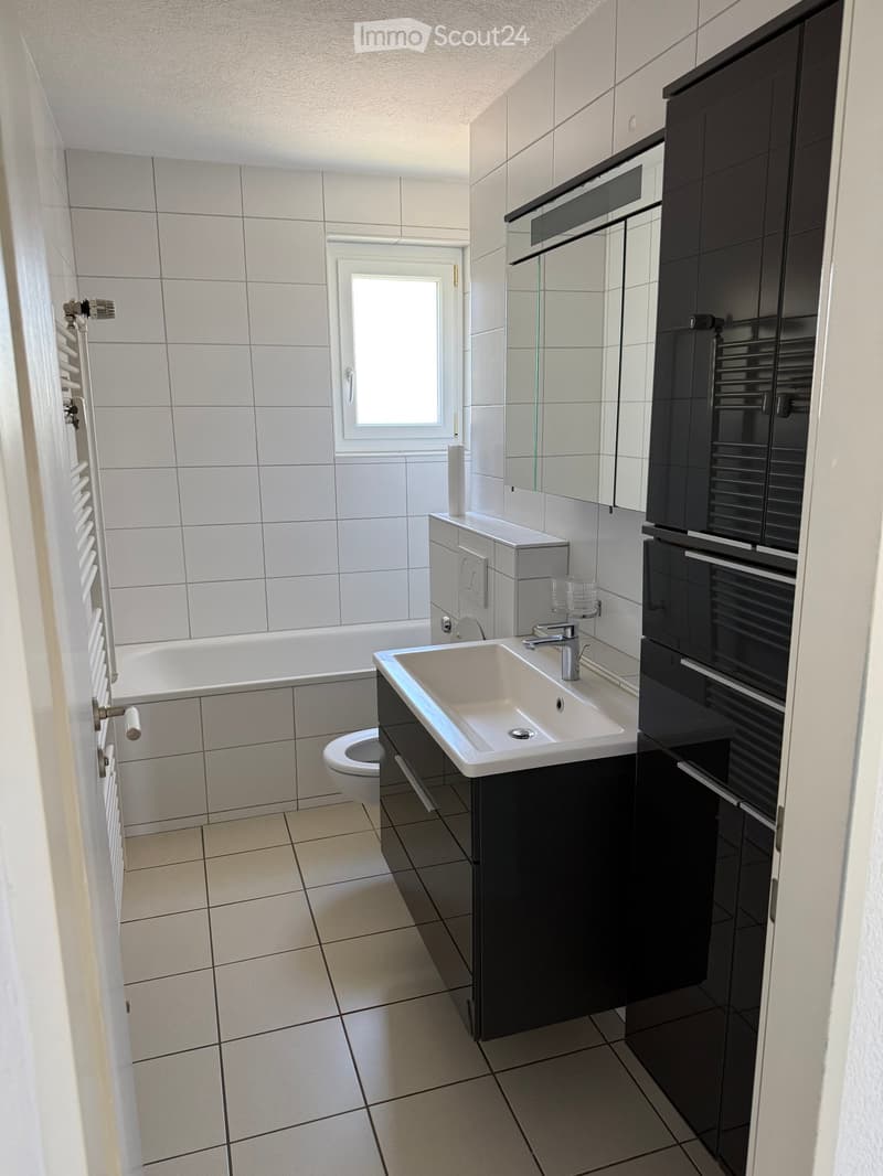 Modern bathroom with white tiles, bathtub, toilet, sink, and dark cabinets.